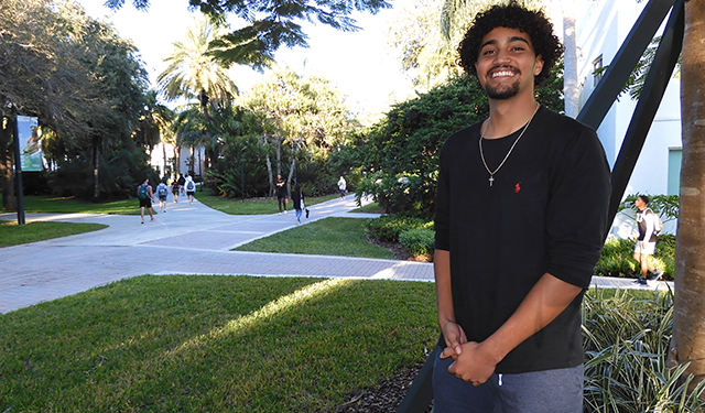 Image of Niko Ferreyra, LAS student, smiling at the camera in front of the UM campus. He is wearing a black t-shirt with a red polo emblem. 